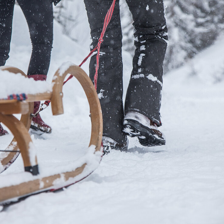 Zwei Menschen in Winterkleidung stehen im Schnee und halten ein Seil fest, das an einer Rodel befestigt ist. Es schneit, und der Schlitten ist teilweise im Schnee vergraben, umgeben von verschneiten Bäumen.