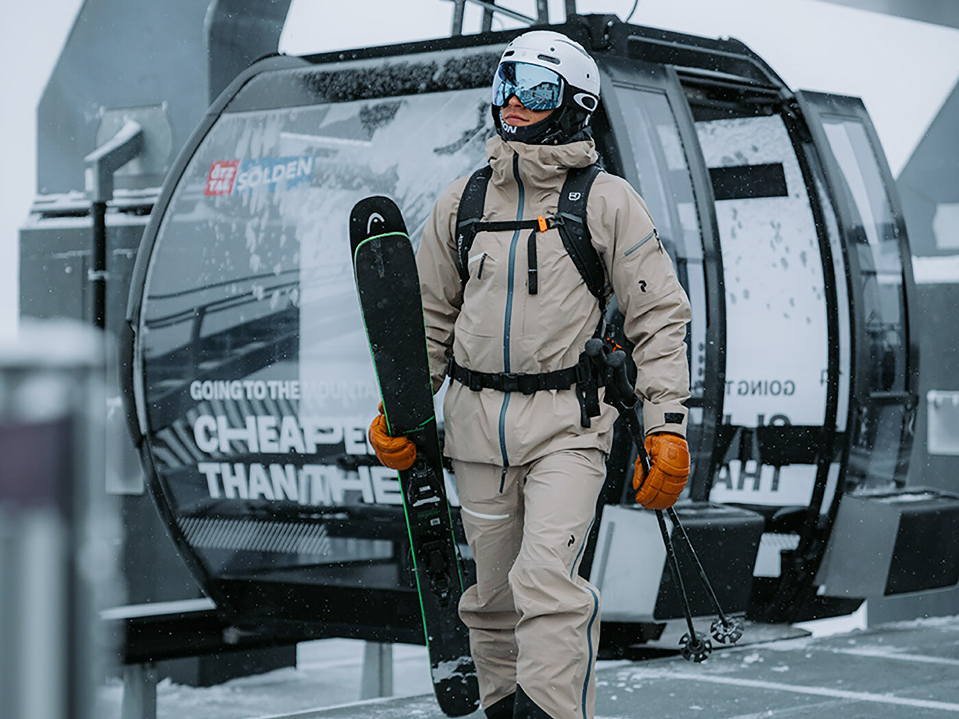Ein Skifahrer in beiger Skikleidung und orangefarbenen Handschuhen trägt seine Skier, während er in einem verschneiten Wintersportort in der Nähe einer Gondelbahn entlanggeht.