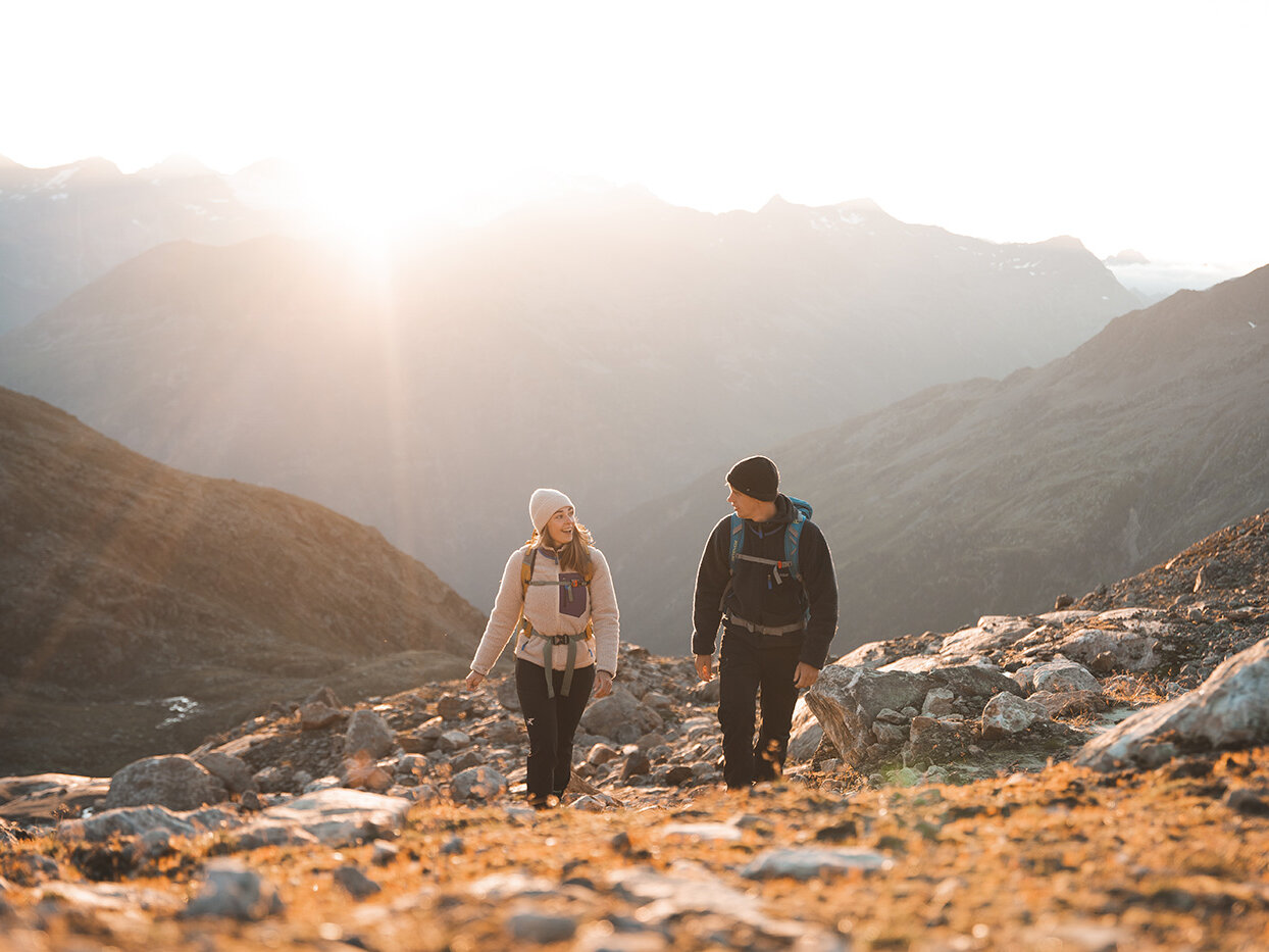 Zwei Menschen mit Rucksäcken und warmer Kleidung wandern bei Sonnenaufgang einen felsigen Bergpfad hinauf, während im Hintergrund das Sonnenlicht über die fernen Berggipfel strahlt.