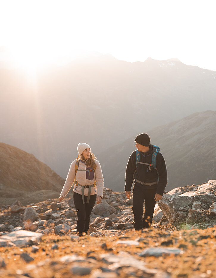 Zwei Menschen mit Rucksäcken und warmer Kleidung wandern bei Sonnenaufgang einen felsigen Bergpfad hinauf, während im Hintergrund das Sonnenlicht über die fernen Berggipfel strahlt.
