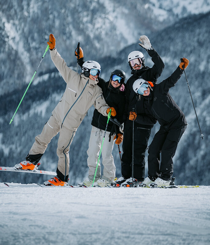 Vier Personen in Skikleidung posieren fröhlich auf einem verschneiten Berghang und halten Skistöcke in den Händen, im Hintergrund sind schneebedeckte Bäume und Berge zu sehen.