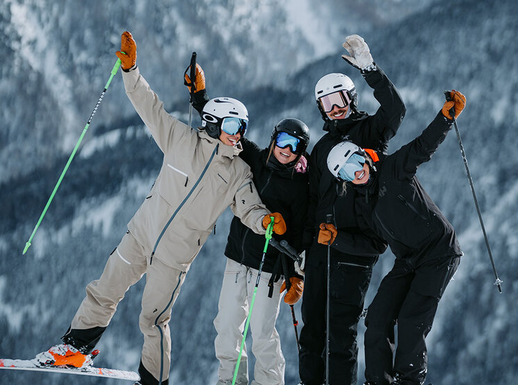 Vier Personen in Skikleidung posieren fröhlich auf einem verschneiten Berghang und halten Skistöcke in den Händen, im Hintergrund sind schneebedeckte Bäume und Berge zu sehen.