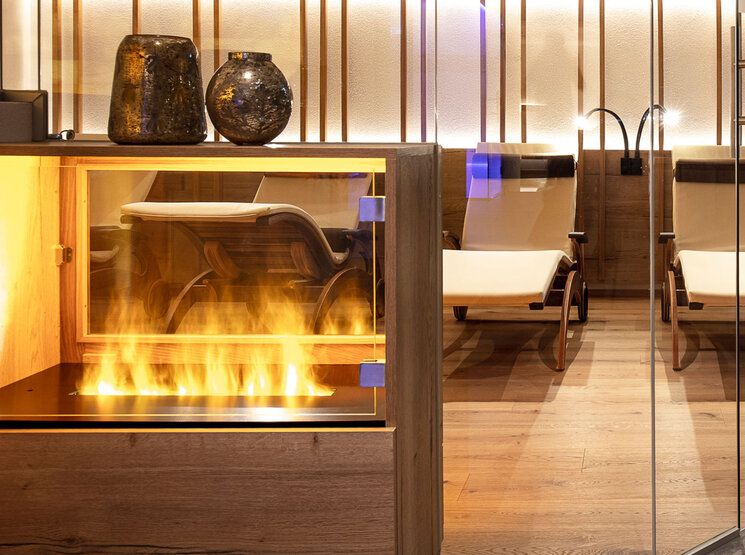 Moderne Spa-Lounge mit Liegestühlen, Holzboden, einem verglasten Zierkamin und zwei Vasen darauf. Warme Beleuchtung sorgt für eine gemütliche, entspannte Atmosphäre.