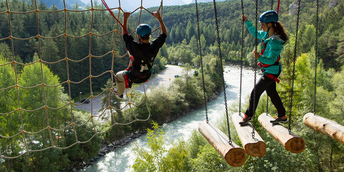 Zwei Personen mit Helmen bewältigen hoch über einem Fluss einen Hochseilgarten in der Area 47: Die eine klettert an einem Seilnetz empor, die andere geht auf hängenden Baumstämmen, umgeben von Bäumen und Bergen.
