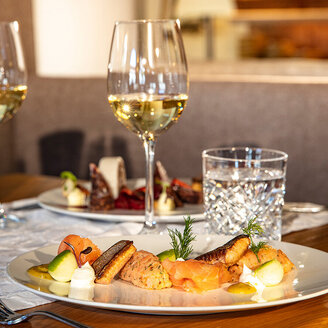 Ein gedeckter Esstisch mit zwei Tellern voller Gourmetgerichte, zwei Gläsern Weißwein und zwei Gläsern Wasser in einem warm beleuchteten Restaurant.