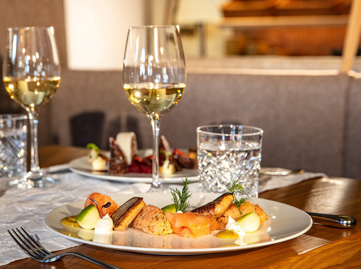 Ein gedeckter Esstisch mit zwei Tellern voller Gourmetgerichte, zwei Gläsern Weißwein und zwei Gläsern Wasser in einem warm beleuchteten Restaurant.