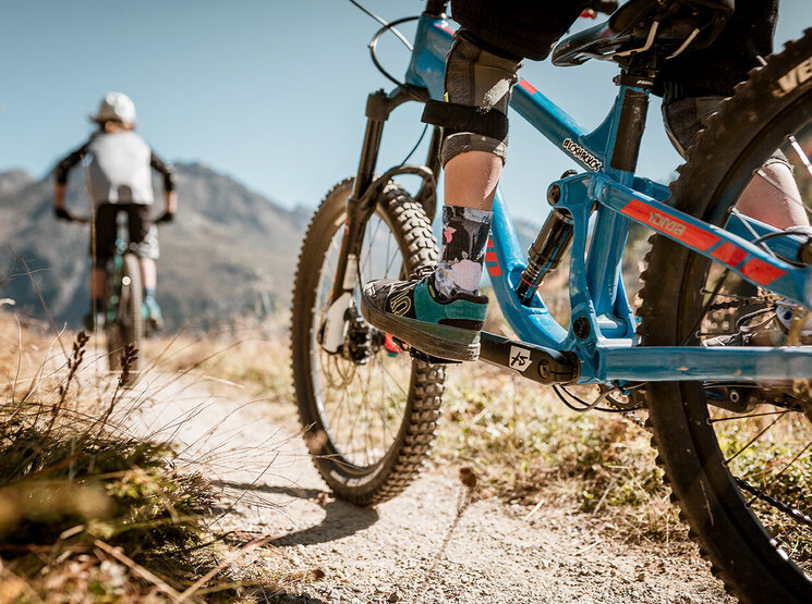 Zwei Radfahrer fahren an einem sonnigen Tag mit Mountainbikes auf einem Feldweg durch grasbewachsenes Gelände, im Hintergrund sind Berge zu sehen; sie tragen Helme und Schutzkleidung.