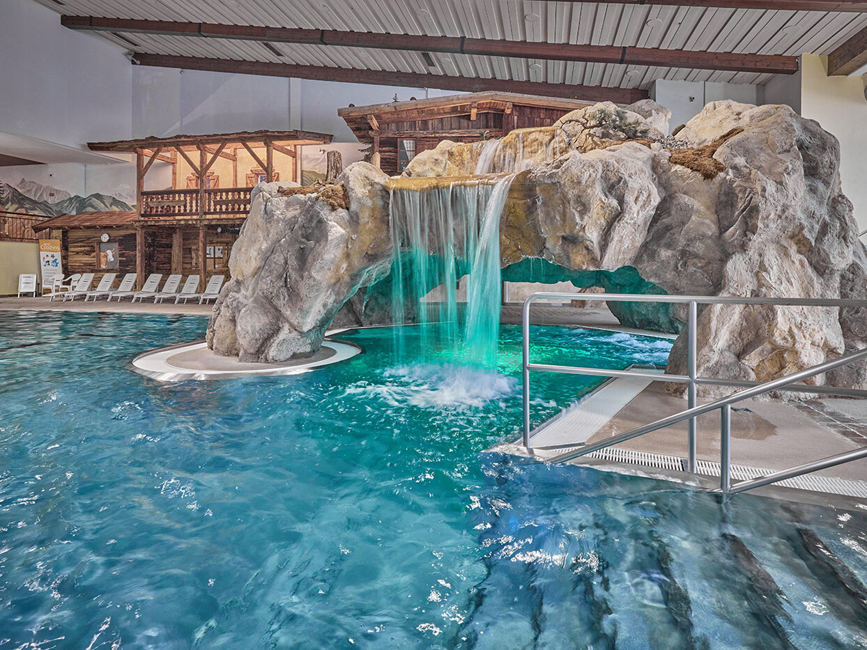 Hallenbad mit einer großen Felsformation, die einen Wasserfall und einen Tunnel umfasst. Unter dem Wasserfall schimmert das Wasser türkis. Im Hintergrund sind Liegestühle am Beckenrand und eine Einrichtung im Chalet-Stil zu sehen.