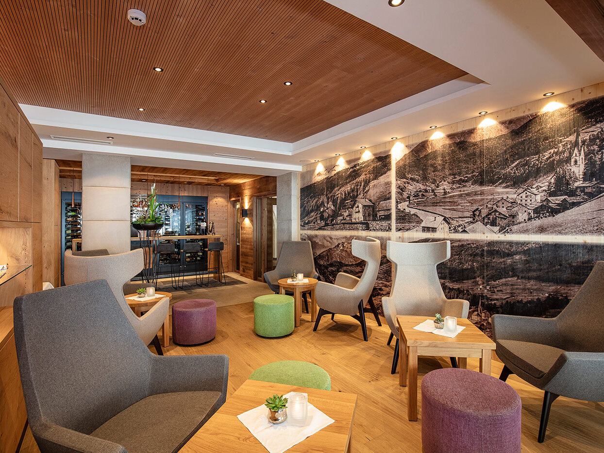 Moderne Lounge mit grauen Sesseln, kleinen Holztischen und bunten Hockern. Eine große Wandmalerei ziert eine der Wände. Der Raum ist warm beleuchtet und mit Holzakzenten versehen, was eine gemütliche, einladende Atmosphäre schafft.