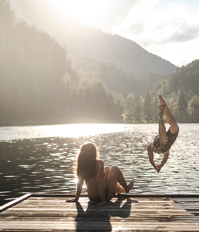 Eine Person sitzt auf einem Holzsteg an einem See, während eine andere Person ins Wasser springt. Im Hintergrund scheint die Sonne über bewaldete Berge.