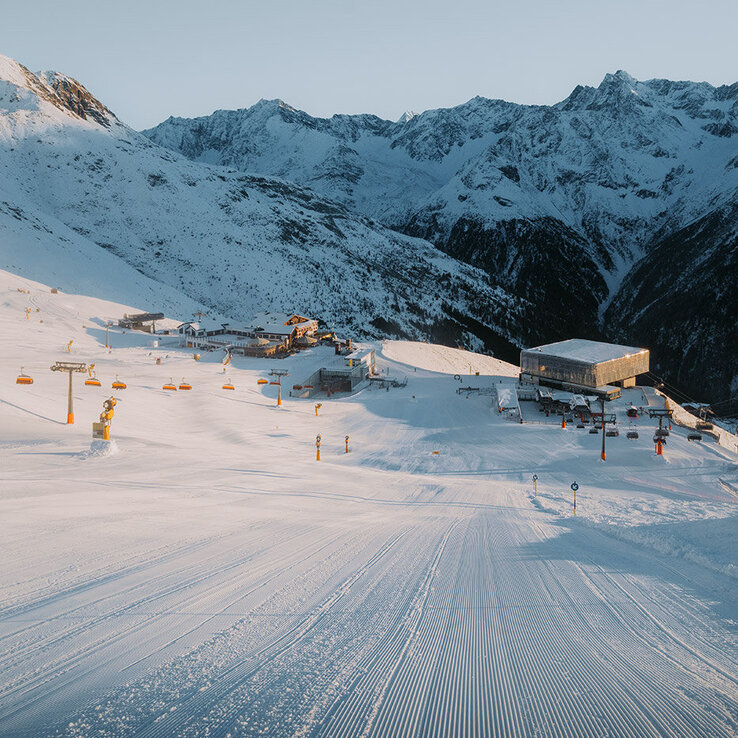 Ein verschneites Skigebiet mit präparierten Pisten, Skiliften und Gebäuden vor schroffen, sonnenbeschienenen Bergen unter einem klaren Himmel.