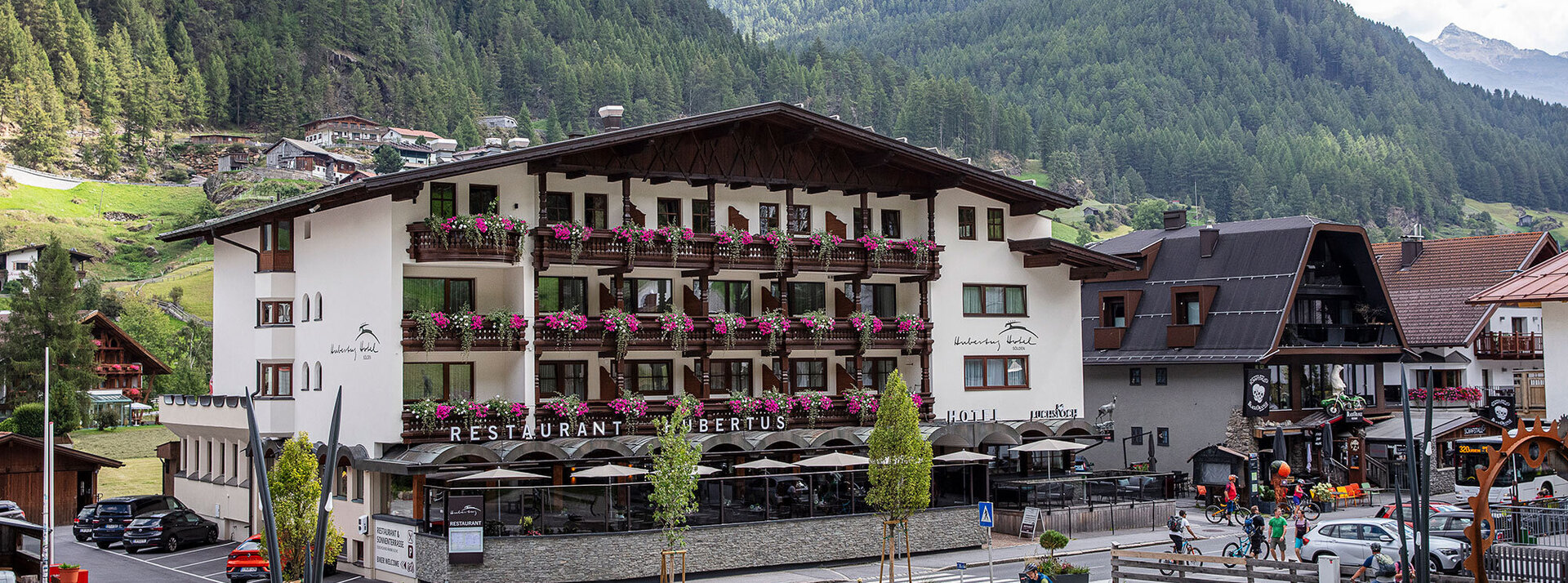Das Hotel Hubertus mit blumengeschmückten Balkonen liegt inmitten von mit Kiefern bewachsenen Bergen; auf der Straße davor sind Radfahrer und Fußgänger unterwegs, während der Himmel teilweise bewölkt ist.