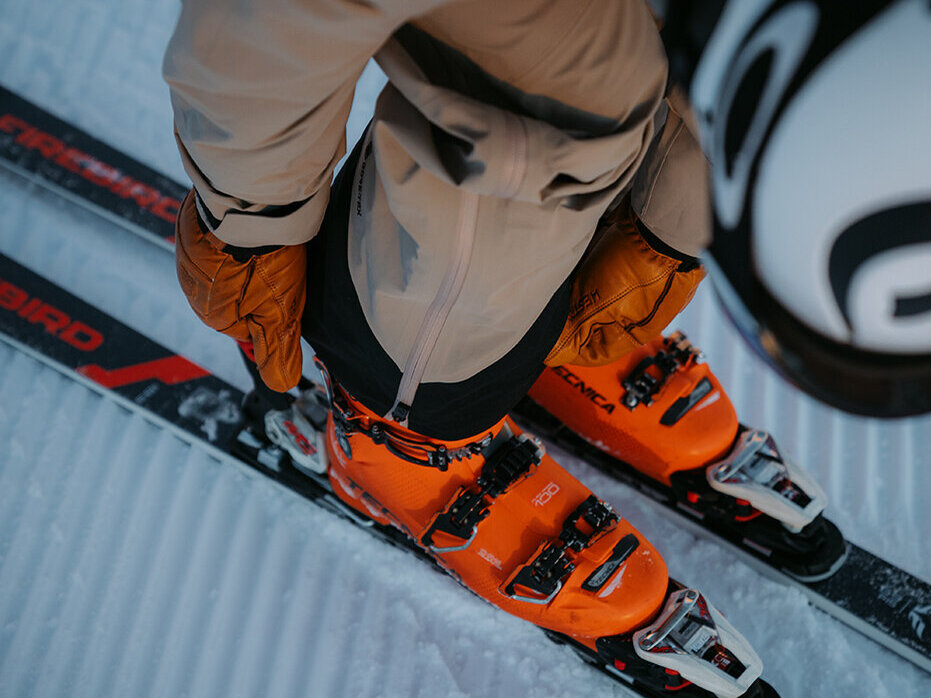 Ein Skifahrer mit orangefarbenen Skischuhen und einem Helm steht auf schwarzen Skiern auf präpariertem Schnee und stellt seine Skibindungen ein.