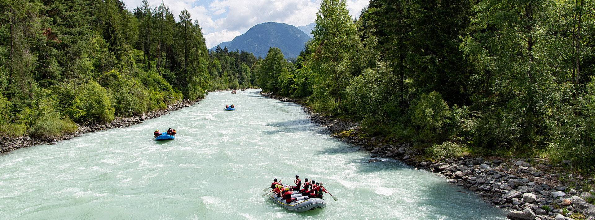 Menschen in Schlauchbooten fahren einen breiten, reißenden Fluss hinunter, umgeben von grünen Bäumen und fernen Bergen unter einem teilweise bewölkten Himmel.