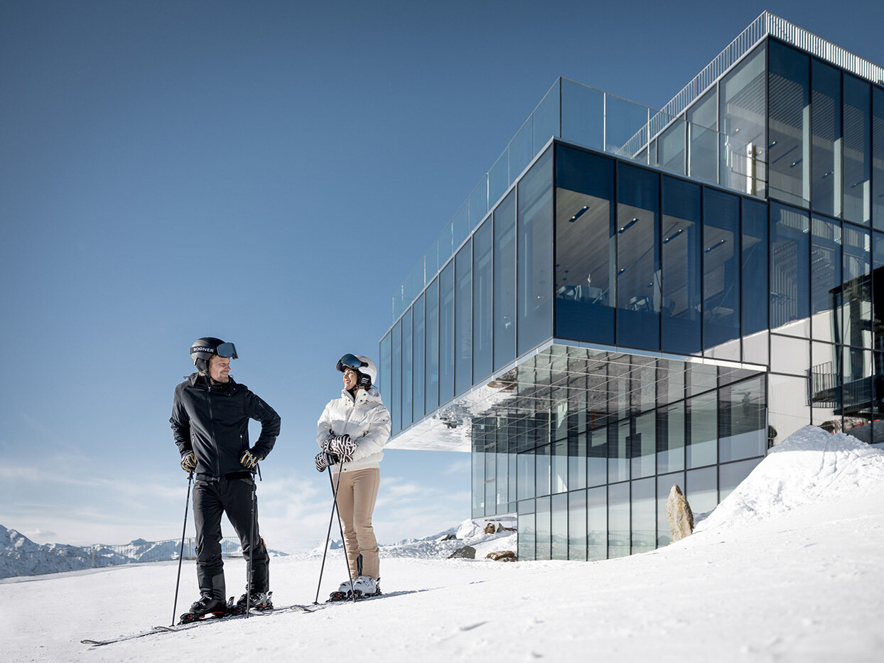 Zwei Personen in Skikleidung stehen auf verschneitem Boden und unterhalten sich vor einem modernen Glasgebäude, während im Hintergrund unter einem klaren Himmel Berge zu sehen sind.