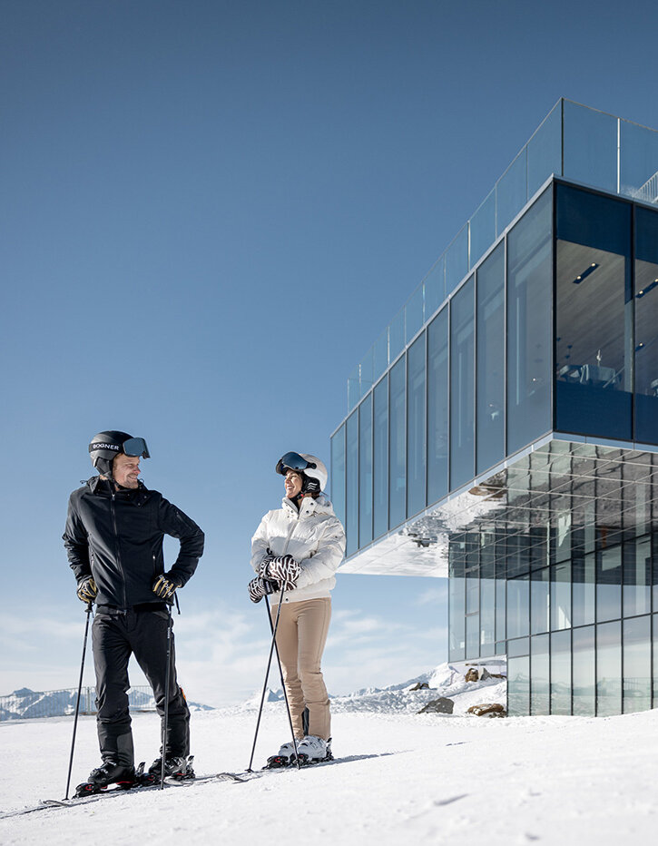 Zwei Personen in Skikleidung stehen auf verschneitem Boden und unterhalten sich vor einem modernen Glasgebäude, während im Hintergrund unter einem klaren Himmel Berge zu sehen sind.