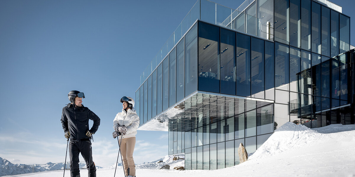 Zwei Personen in Skikleidung stehen auf verschneitem Boden und unterhalten sich vor einem modernen Glasgebäude, während im Hintergrund unter einem klaren Himmel Berge zu sehen sind.