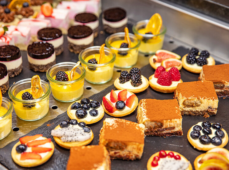 Eine Auswahl an Desserts, darunter Obsttörtchen, Tiramisu-Quadrate, Mini-Käsekuchen und Mousse-Törtchen, garniert mit frischen Früchten und Schokolade, präsentiert auf einer schwarzen Platte.