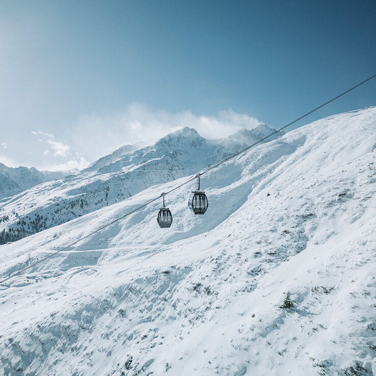 Zwei Seilbahnkabinen gleiten an einem Seil entlang über eine verschneite Berglandschaft unter einem strahlend blauen Himmel.