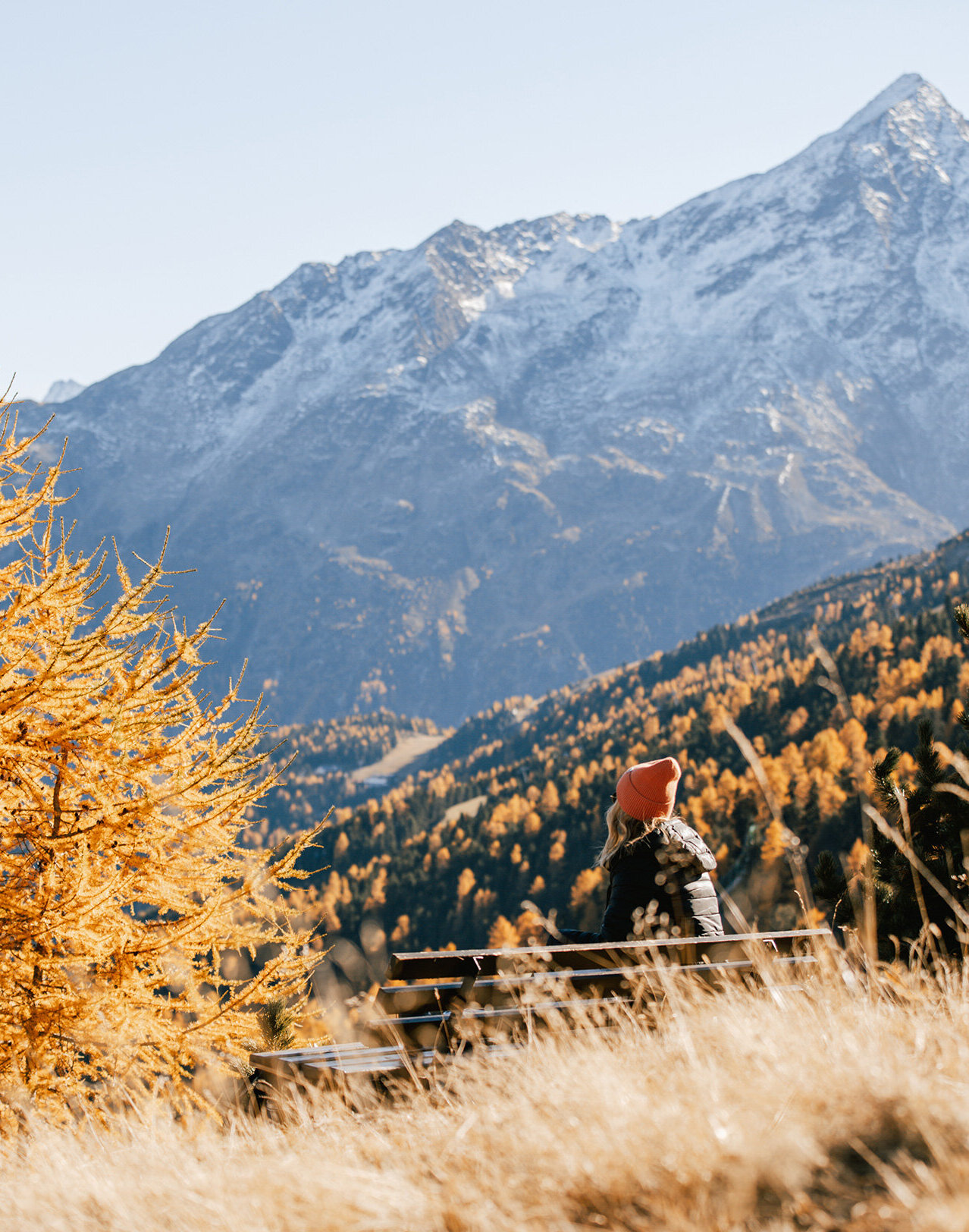 Eine Person mit einer orangefarbenen Mütze sitzt auf einer Bank mit Blick auf schneebedeckte Berge, umgeben von goldfarbenen Herbstbäumen und trockenem Gras unter einem klaren Himmel.