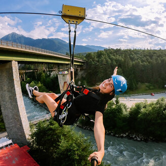Eine lächelnde Person mit einem blauen Helm gleitet an einem sonnigen Tag auf einer Seilrutsche über einen Fluss, umgeben von Bäumen und Bergen, im Hintergrund ist eine Brücke zu sehen.