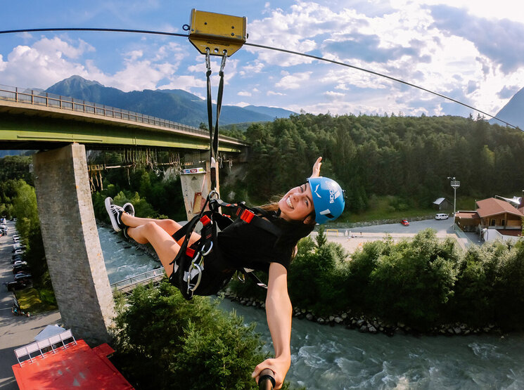 Eine lächelnde Person mit einem blauen Helm gleitet an einem sonnigen Tag auf einer Seilrutsche über einen Fluss, umgeben von Bäumen und Bergen, im Hintergrund ist eine Brücke zu sehen.