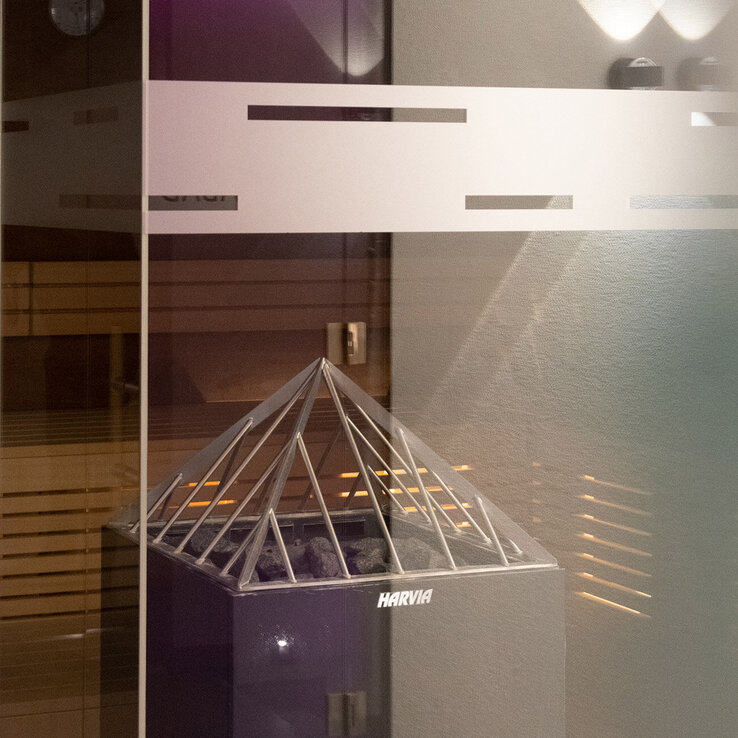 Ein moderner Saunaofen mit einem pyramidenförmigen Metallrahmen oben ist in einem Saunaraum aus Holz teilweise durch eine Glastür mit mattierten horizontalen Linien zu sehen.