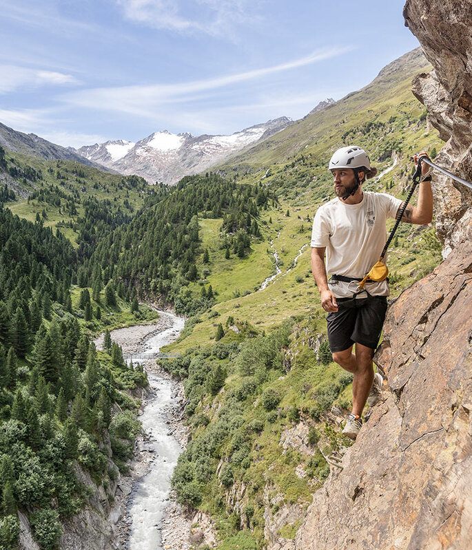 Eine Person mit Helm erklimmt einen felsigen Bergpfad, der mit Seilen gesichert ist, und blickt auf ein bewaldetes Tal mit einem sich schlängelnden Fluss und fernen schneebedeckten Gipfeln unter blauem Himmel.