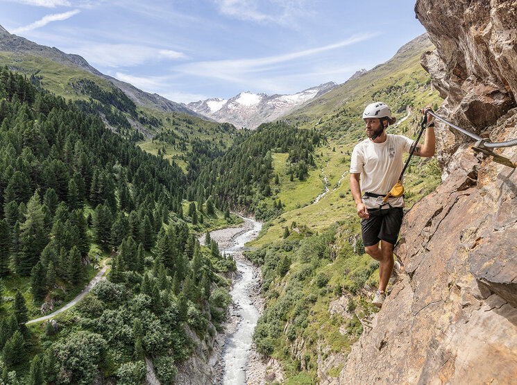 Eine Person mit Helm erklimmt einen felsigen Bergpfad, der mit Seilen gesichert ist, und blickt auf ein bewaldetes Tal mit einem sich schlängelnden Fluss und fernen schneebedeckten Gipfeln unter blauem Himmel.