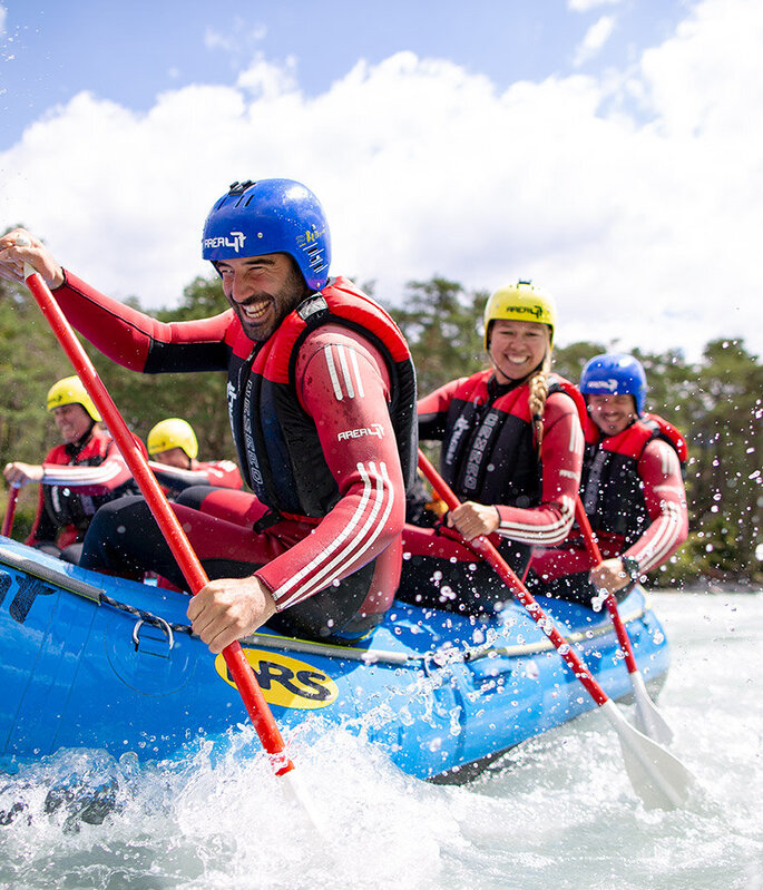 Eine Gruppe von Menschen, die Helme und Schwimmwesten tragen, paddelt gemeinsam in einem blauen Schlauchboot auf einem Fluss und spritzt dabei Wasser in die Luft, während sie das Wildwasser-Rafting genießen. Im Hintergrund sind Bäume und Berge zu sehen.