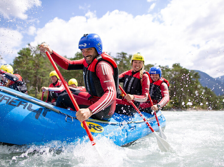 Eine Gruppe von Menschen, die Helme und Schwimmwesten tragen, paddelt gemeinsam in einem blauen Schlauchboot auf einem Fluss und spritzt dabei Wasser in die Luft, während sie das Wildwasser-Rafting genießen. Im Hintergrund sind Bäume und Berge zu sehen.