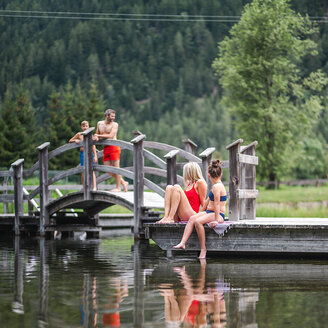 Zwei Frauen sitzen auf einem hölzernen Steg am Wasser, während im Hintergrund zwei Männer auf einer kleinen Brücke stehen und sich unterhalten, umgeben von grünen Bäumen und Bergen.