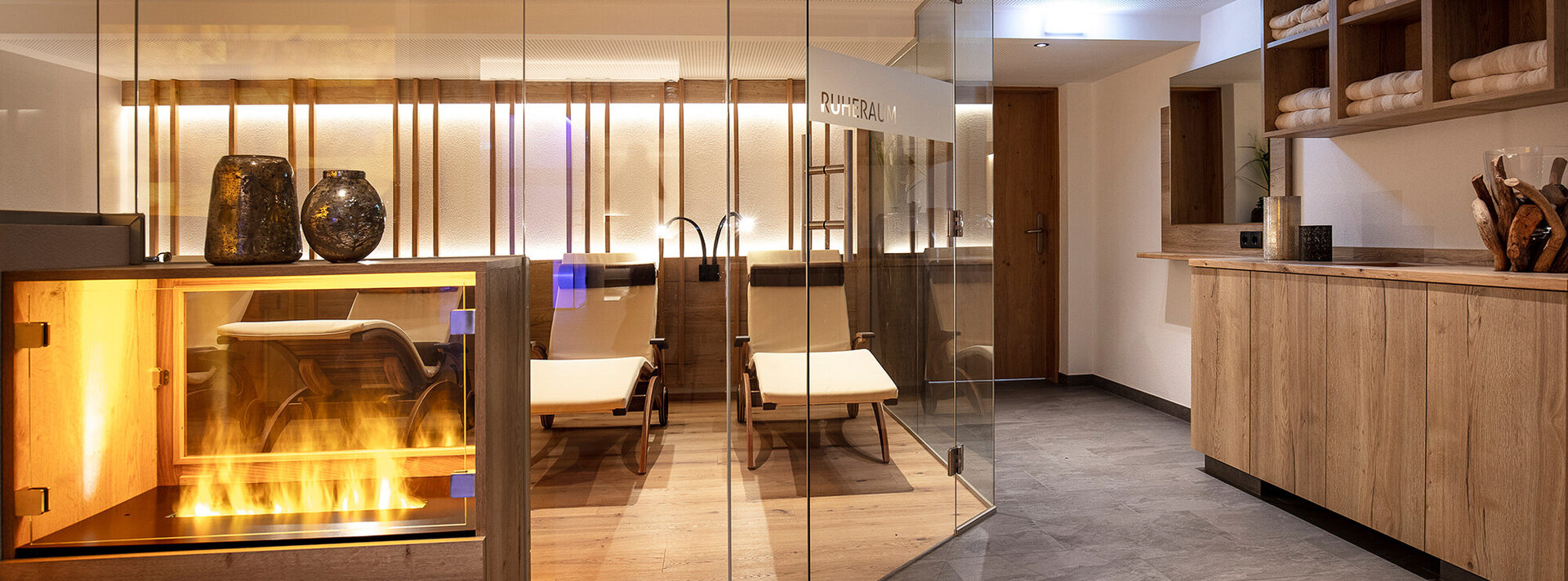 Ein moderner Wellness-Ruheraum mit Glaswänden, Liegestühlen, ordentlich auf Regalen gestapelten Handtüchern und einem prasselnden Kamin neben dem Eingang. Die Einrichtung besticht durch warme Holztöne und sanfte Beleuchtung.