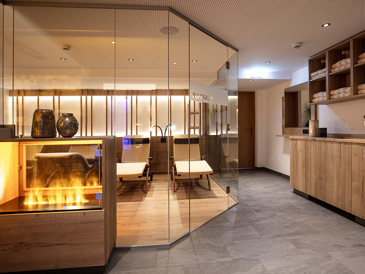 Moderne Spa-Lounge mit einem verglasten Ruheraum, Liegen, einem Kamin, Regalen mit aufgerollten Handtüchern und warmer Stimmungsbeleuchtung.