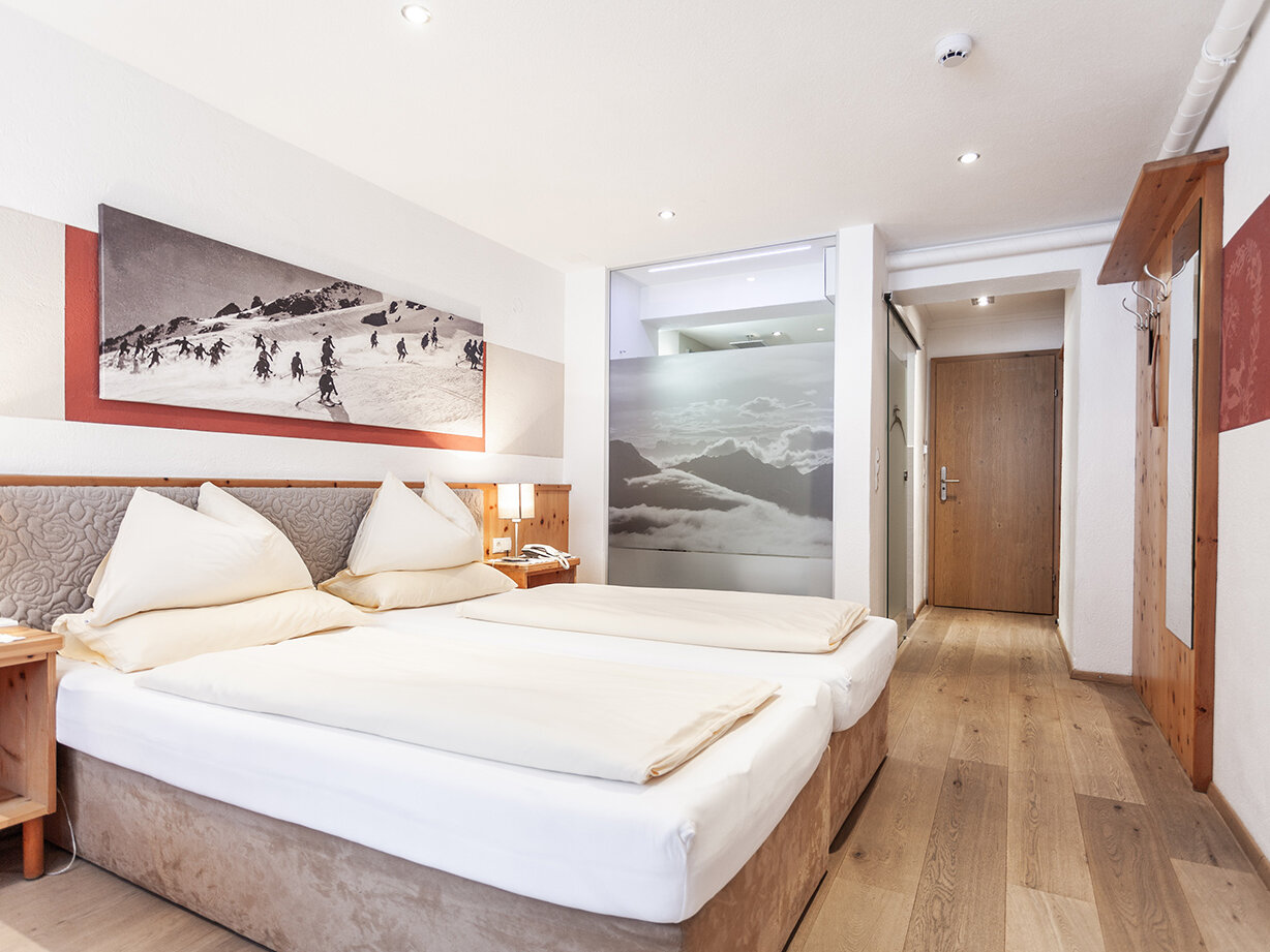 Ein helles Hotelzimmer mit zwei aneinandergeschobenen Einzelbetten, Holzmöbeln, einem Wandbild mit Bergmotiv über dem Kopfteil und einem Holzboden. Eine offene Tür führt in einen Flur mit Kunstwerken aus Milchglas.
