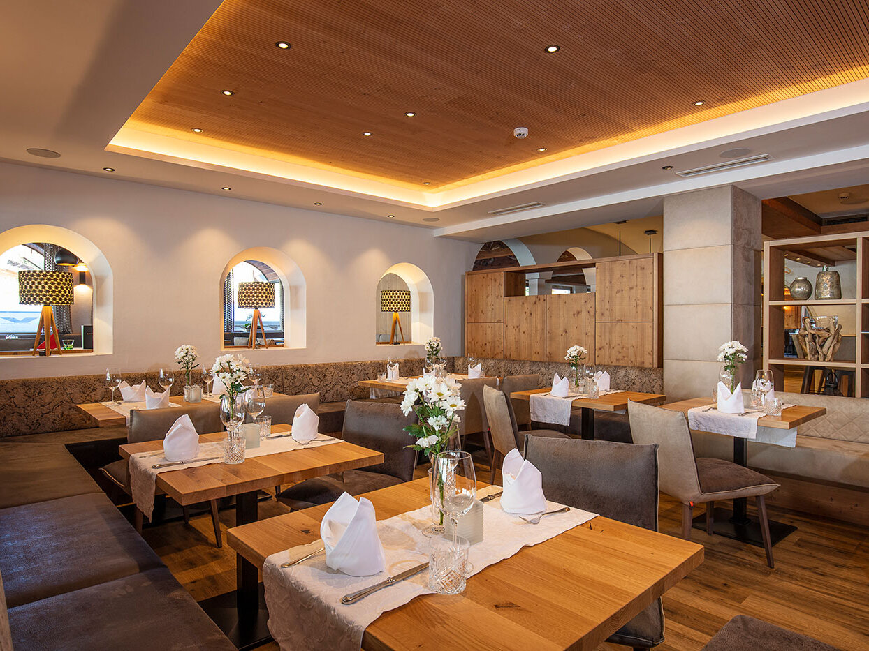 Ein gemütliches Restaurantinterieur mit Holztischen, gepolsterten Sitzbänken, ordentlich gefalteten Servietten, weißen Blumen in Vasen, sanfter Beleuchtung sowie dekorativen Regalen und Spiegeln an den Wänden.