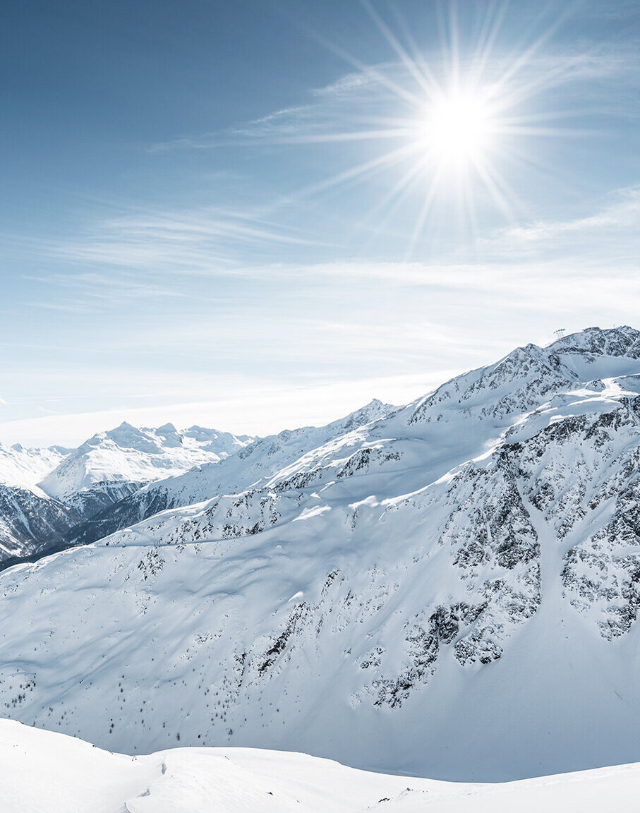 Schneebedeckte Berggipfel unter strahlendem Sonnenschein und klarem blauem Himmel, über dem sich leichte Wolken kaum erkennen lassen. Das Sonnenlicht spiegelt sich im weißen Schnee wider und schafft so eine friedliche Winterlandschaft.