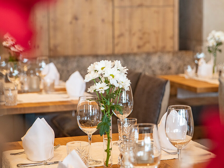 Ein Holztisch in einem gemütlichen Restaurant ist mit weißen Servietten, Gläsern und einer Vase mit weißen Blumen gedeckt und erwartet die Gäste. Unscharfe rote Blumen rahmen den Vordergrund ein.