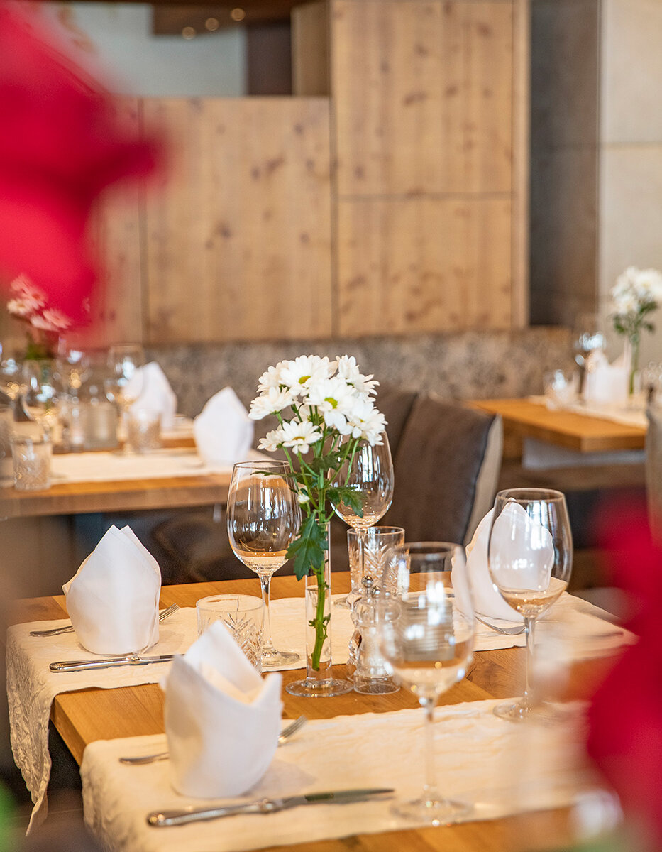 Ein Holztisch in einem gemütlichen Restaurant ist mit weißen Servietten, Gläsern und einer Vase mit weißen Blumen gedeckt und erwartet die Gäste. Unscharfe rote Blumen rahmen den Vordergrund ein.
