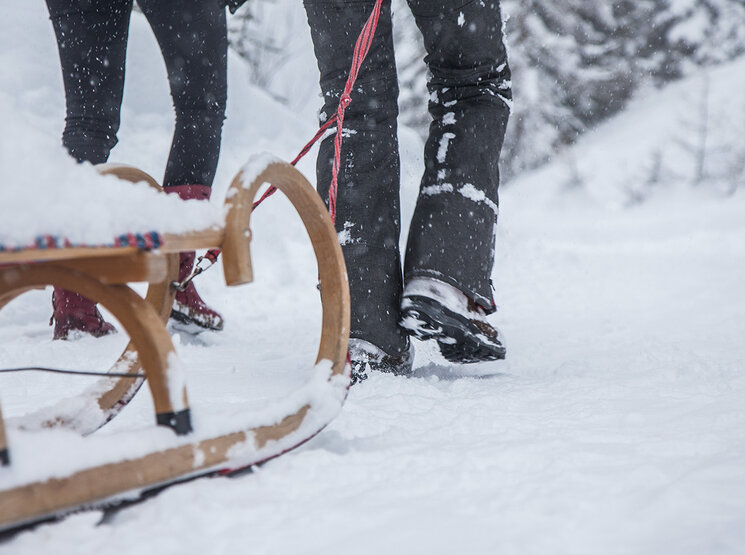 Zwei Menschen stehen im Schnee neben einem Holzschlitten. Schnee bedeckt den Boden und den Schlitten, im verschneiten Hintergrund sind Bäume zu sehen. Nur ihre Beine und Füße sind zu sehen.