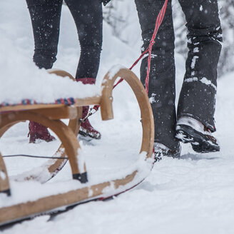 Zwei Menschen stehen im Schnee neben einem Holzschlitten. Schnee bedeckt den Boden und den Schlitten, im verschneiten Hintergrund sind Bäume zu sehen. Nur ihre Beine und Füße sind zu sehen.