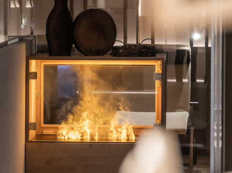 Ein moderner Elektrokamin mit realistisch wirkenden Flammen ist in ein Holzmöbelstück eingebaut. Darauf stehen zwei dekorative Vasen, im Hintergrund ist ein unscharf abgebildetes, modernes Interieur zu sehen.