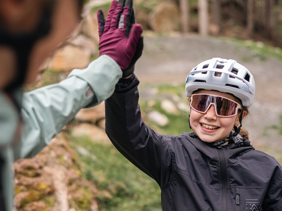 Zwei Radfahrer mit Helm und Sonnenbrille geben sich im Freien auf einem Waldweg ein High-Five. Eine Person lächelt breit, während die andere im Vordergrund teilweise unscharf zu sehen ist.