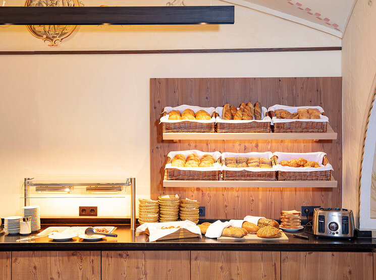 In einem Holzregal über der Theke sind verschiedene Brotsorten und Backwaren in Körben ausgestellt; auf der Theke stehen gestapelte Teller, ein Toaster und Tassen in einem warm beleuchteten Frühstücksbereich.