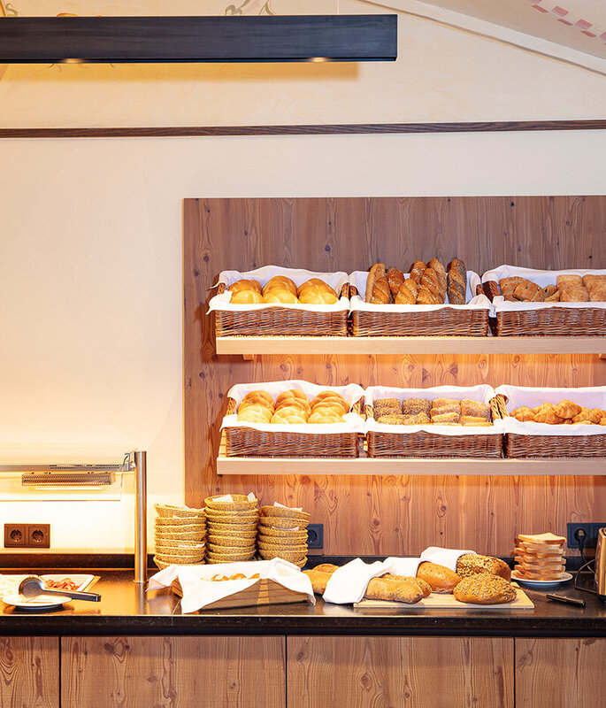 In einem Holzregal über der Theke sind verschiedene Brotsorten und Backwaren in Körben ausgestellt; auf der Theke stehen gestapelte Teller, ein Toaster und Tassen in einem warm beleuchteten Frühstücksbereich.