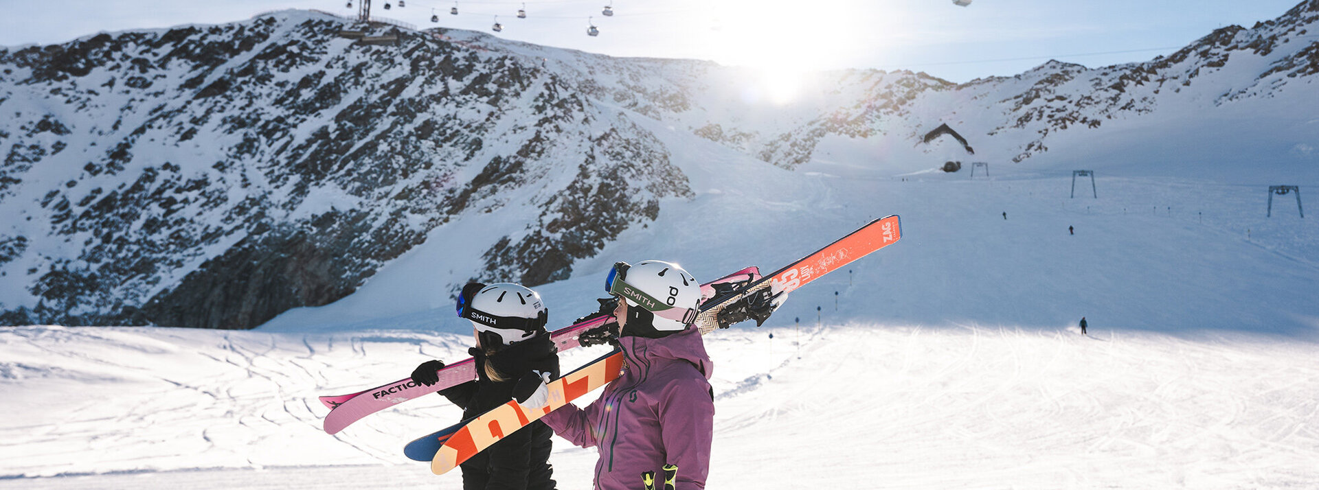 Zwei Skifahrer mit Helmen und Winterbekleidung tragen ihre Skier auf den Schultern und gehen einen verschneiten Berghang hinauf, im Hintergrund sind Skilifte und Sonnenlicht zu sehen.