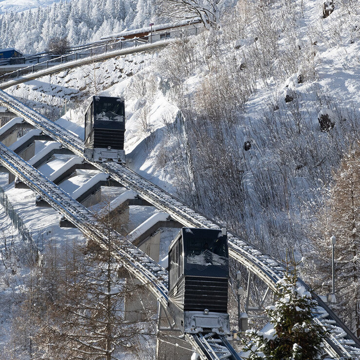 Zwei Standseilbahnwagen fahren auf parallelen Gleisen unter einem strahlend blauen Himmel einen verschneiten, bewaldeten Berghang hinauf, im Hintergrund sind schneebedeckte Nadelbäume und ferne Berggipfel zu sehen.