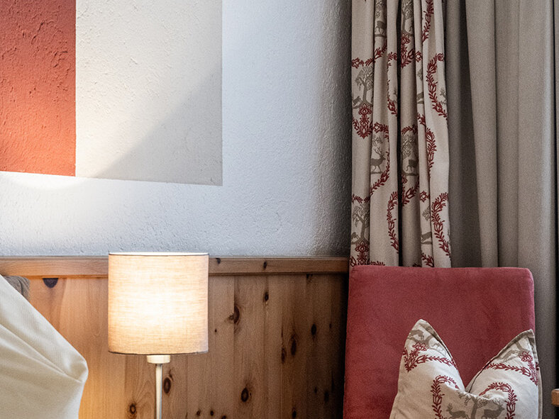 Gemütliche Schlafzimmer-Ecke mit einem ordentlich gemachten Bett, einem rot-weißen Zierkissen, einem Nachttisch aus Holz, einer brennenden Lampe, gemusterten Vorhängen und einem abstrakten Wandbild über dem Kopfteil.