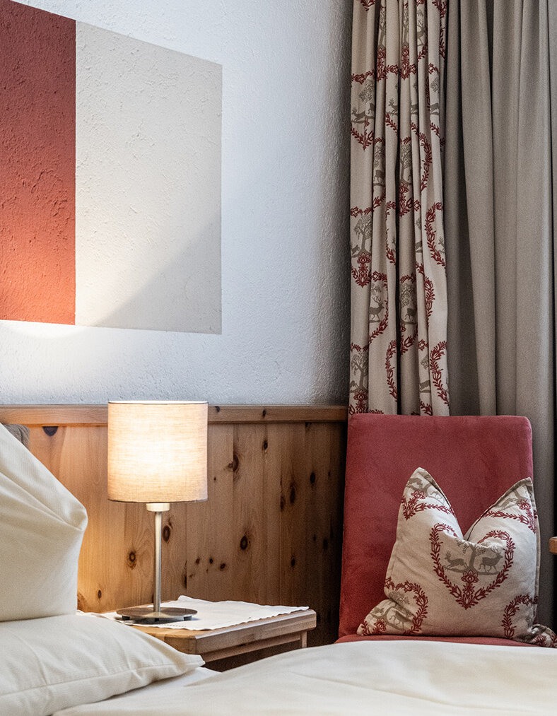 Gemütliche Schlafzimmer-Ecke mit einem ordentlich gemachten Bett, einem rot-weißen Zierkissen, einem Nachttisch aus Holz, einer brennenden Lampe, gemusterten Vorhängen und einem abstrakten Wandbild über dem Kopfteil.