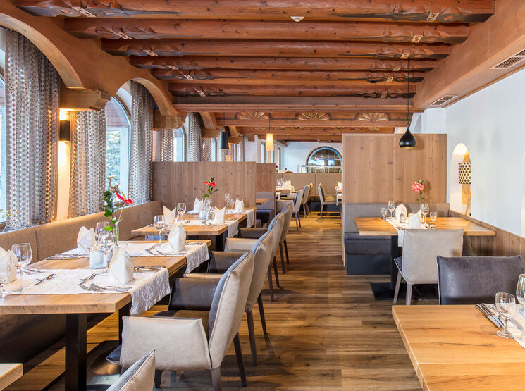Ein elegantes Restaurant mit Holzbalken, großen Bogenfenstern, gepflegt gedeckten Tischen und gepolsterten Stühlen. Der Raum ist von natürlichem Licht durchflutet, und auf jedem Tisch stehen Blumen, was eine warme und einladende Atmosphäre schafft.